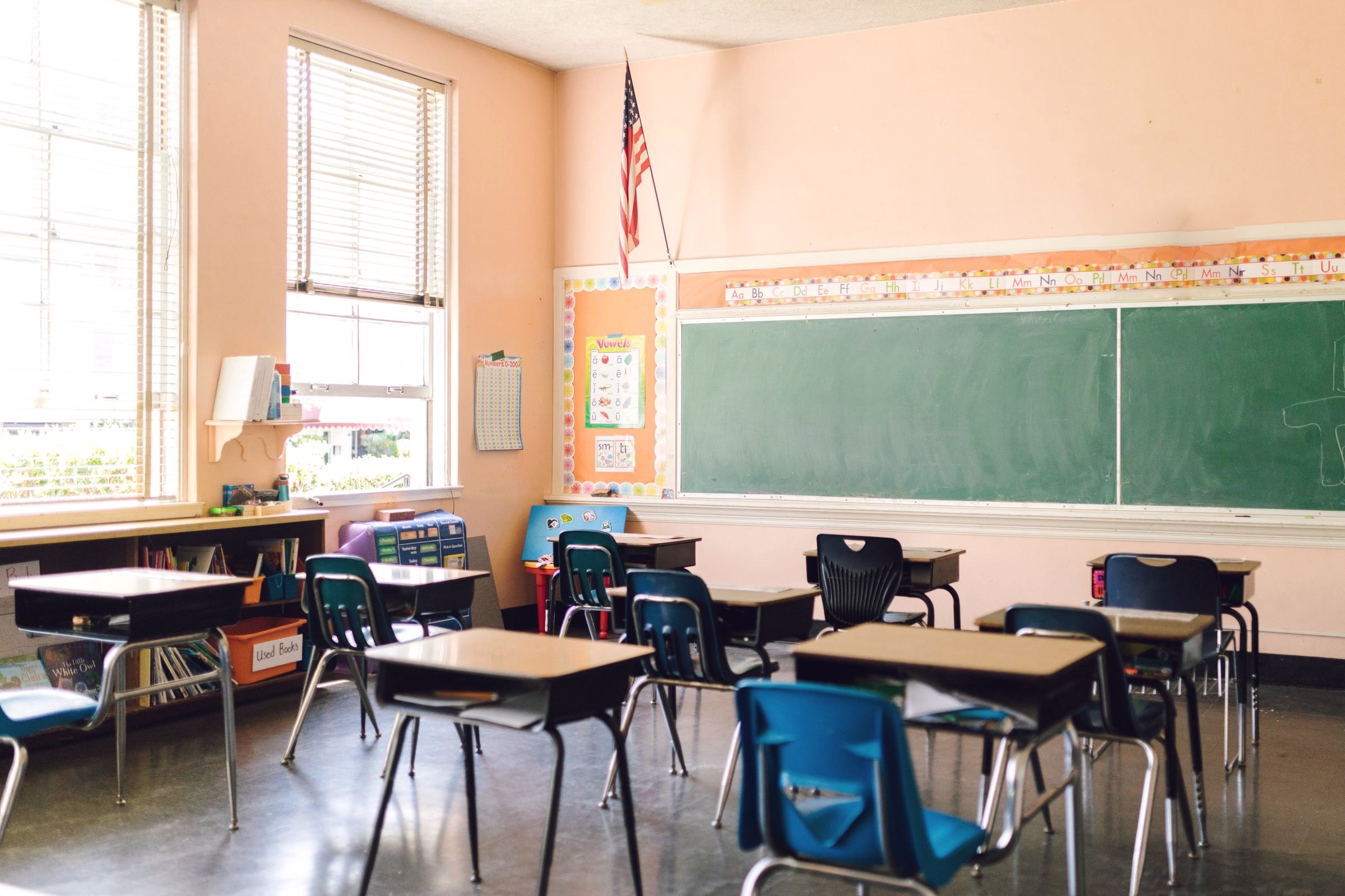 An empty, sunlit elementary classroom with desks in rows and a flag by the window.