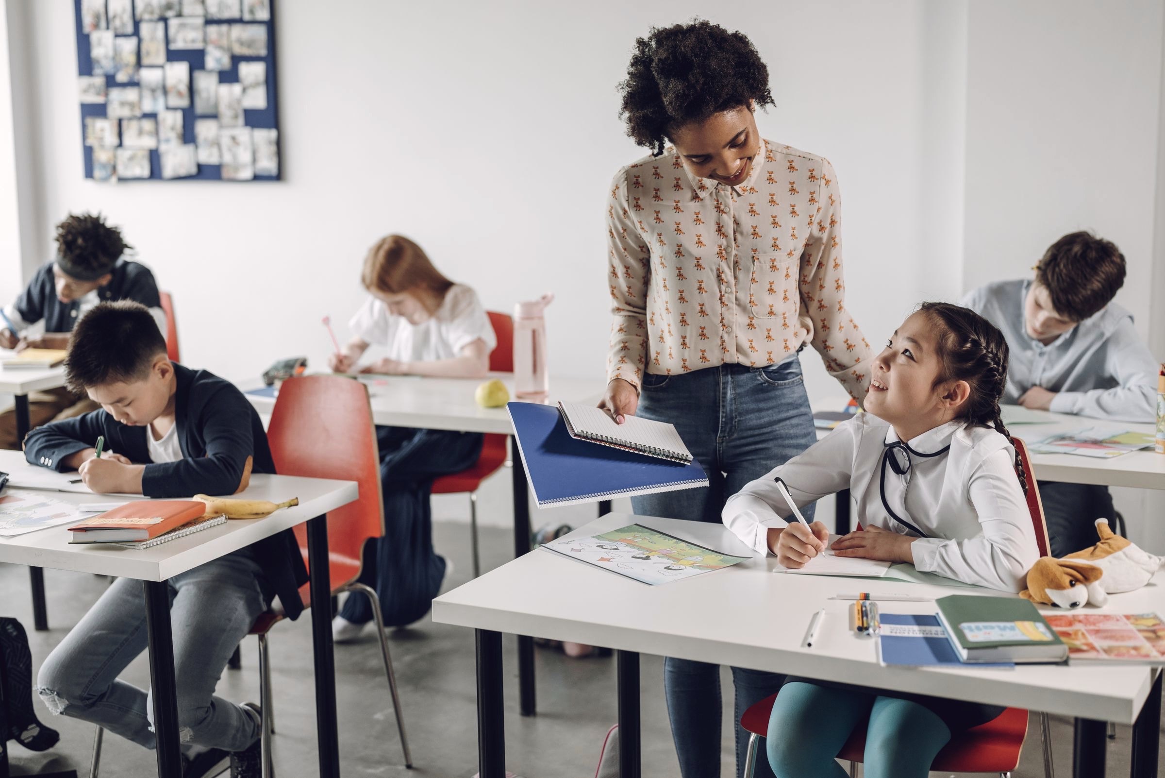A teacher handing back work to a student at her desk while the class writes